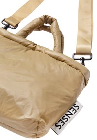 Beige padded bag with a textured surface, featuring a top handle and an adjustable strap. Includes a visible brand tag.