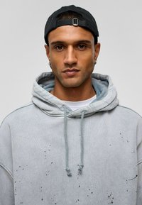 Lichtgrijze oversized hoodie met een voorzak, koord en zwarte verfspatten details. Gedragen onder een zwarte pet.