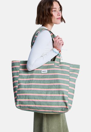 Jonge vrouw in een wit shirt met lange mouwen draagt een grote draagtas met groene, roze en gele strepen met het label "ROXY" op de schouder.