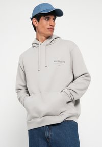 Sweat à capuche gris avec une poche avant et un cordon de serrage, arborant le logo "AllSaints". Associé à un jean en denim bleu et une casquette bleue.