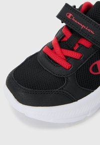 Chaussure de sport noire avec une tige en mesh texturé, accents rouges sur les lacets et la marque, bande Velcro, et une semelle en caoutchouc blanche.
