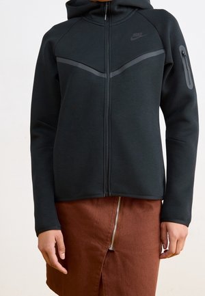 Czarna bluza Nike na zamek z odblaskowymi paskami i kieszenią na zamek na rękawie, noszona z brązową spódnicą z zamkiem z przodu.