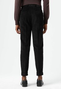 Zwarte corduroy broek met een nauwsluitende pasvorm, voorzien van twee achterzakken, opgerolde manchetten en een gestructureerd oppervlak. Gesloten met zwarte schoenen.