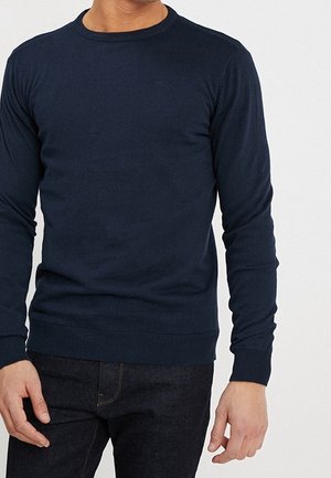 Mann trägt einen enganliegenden, langärmeligen, dunkelblauen Pullover mit Rundhalsausschnitt und dunkle Jeans vor einem schlichten hellen Hintergrund.