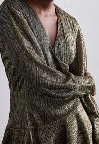 Robe métallisée dorée avec un décolleté en V profond, des manches longues et un ourlet évasé. Texturée avec de fines rayures verticales, créant un effet brillant.