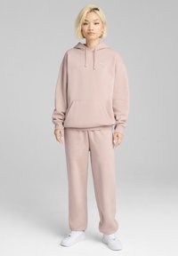 Lyserød hoodie med kængurulomme, matchet med lyserøde sweatpants. Har blødt stof, løs pasform og et lille logo på øverste venstre side. Hvide sneakers.