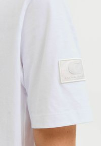 Valge T-särk pehmest puuvillast lühikeste varrukatega. Ülemisel varrukal on tekstuuritud valge patch koos kõrgendatud logoga. Puhtne ja lihtne disain.