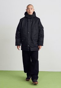 Schwarze gepolsterte Parka mit Kapuze, vorderer Knopfverschluss und zwei großen Seitentaschen, getragen über lockeren schwarzen Hosen und dunklem Schuhwerk.