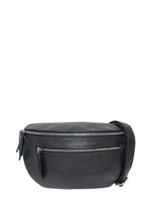 Junge Frau mit langen braunen Haaren, die ein schwarzes Langarmshirt und Jeans trägt und eine schwarze Leder-Umhängetasche mit silbernen Reißverschlüssen hält.