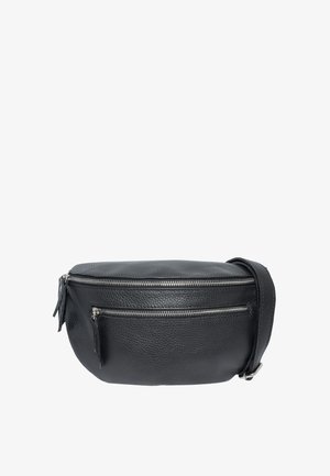 Junge Frau mit langen braunen Haaren, die ein schwarzes Langarmshirt und Jeans trägt und eine schwarze Leder-Umhängetasche mit silbernen Reißverschlüssen hält.