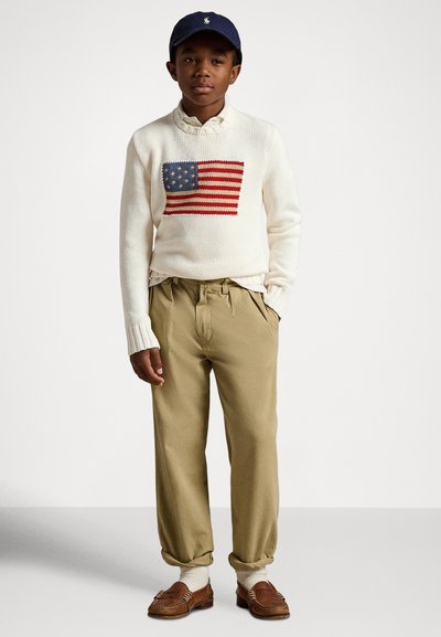 Pleteni krem pulover z vezeno ameriško zastavo, kaki hlače z gubami, svetlobejž nogavice in rjavi loaferji; na glavi mornarska kapa.