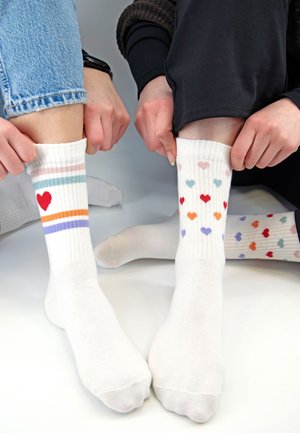 Zwei Personen ziehen bunte weiße Socken mit Herzmuster hoch, bei einem mit Streifen und beim anderen mit mehrfarbigen Herzen an den Beinen.