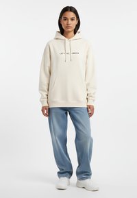 Felpa crema con testo "LET'S GET MARRIED", tasca a marsupio, cappuccio con cordino, abbinata a jeans a gamba larga blu chiaro e sneakers bianche.