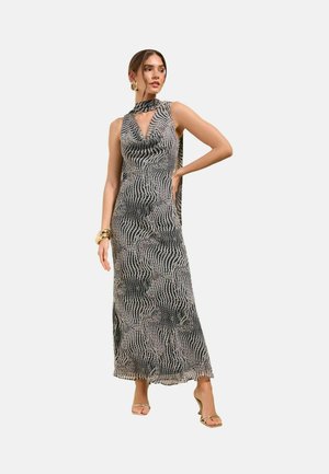 Donna che indossa un abito lungo senza maniche con motivo bianco e nero, collo alto e sciarpa abbinata, accessoriato con braccialetti dorati e sandali.