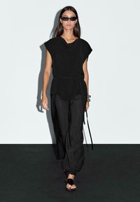Top nero oversize con scollatura drappeggiata, abbinato a pantaloni larghi neri e sandali neri. Il modello indossa occhiali da sole stilosi e stretti.