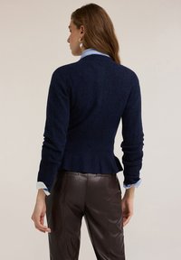 Granatowy sweter z dzianiny o dopasowanej talii i fasonie z baskinką. Posiada długie rękawy oraz warstwowy dół, zestawiony z brązowymi skórzanymi spodniami.