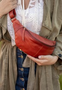 Marsupio in pelle rossa con forma curva, zip superiore e tracolla staccabile, tenuto da una persona che indossa una blusa chiara e una giacca oliva.