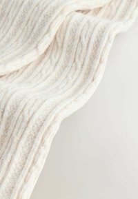 Vitt stickat tyg med ett strukturerat ribbmönster, som visar variationer i tjocklek och en mjuk, fluffig finish. Den böjda draperingen förmedlar en känsla av flöde.