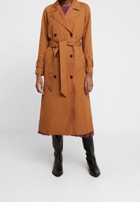 Abrigo trench marrón con botones cruzados, cintura con cinturón y mangas largas; de longitud hasta la rodilla. Llevado sobre un vestido estampado floral y botas negras.