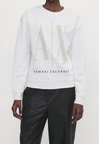 Sudadera blanca con logo "AX" adornado con tachuelas doradas y texto "ARMANI EXCHANGE". Presenta cuello redondo y puños acanalados, combinada con pantalones negros.