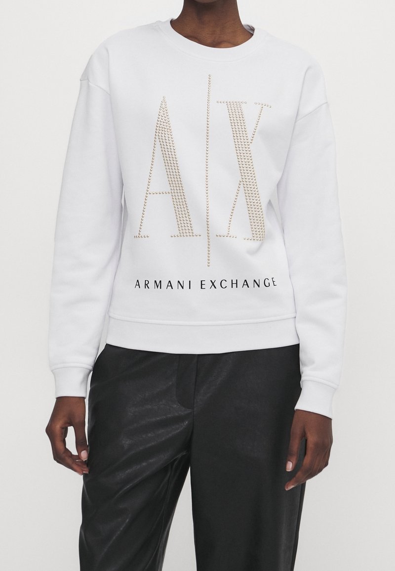Sudadera blanca con logo "AX" adornado con tachuelas doradas y texto "ARMANI EXCHANGE". Presenta cuello redondo y puños acanalados, combinada con pantalones negros.