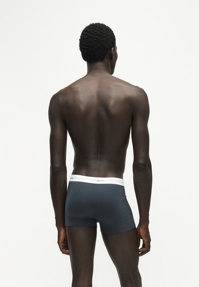Homme à la peau foncée et aux cheveux courts bouclés, debout de dos, portant un boxer bleu foncé avec une ceinture blanche sur un fond uni.