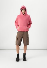 Sweatshirt à capuche rose avec poche kangourou, associé à un short cargo marron avec poches latérales. Des bottes noires à lacets complètent la tenue.