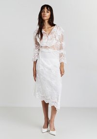 Weißes Spitzenkleid, das über einem satinartigen Unterkleid liegt, mit einem rüschigen, transparenten Überzug und einem gewellten Saum. Spitz zulaufende, weiße Absätze vervollständigen den Look.
