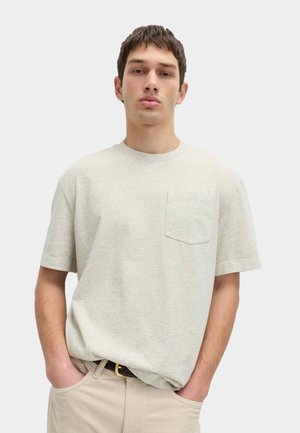 Ung mand med kort mørkt hår iført en lys beige T-shirt med brystlomme og beige bukser, hænderne i lommerne, neutral baggrund.