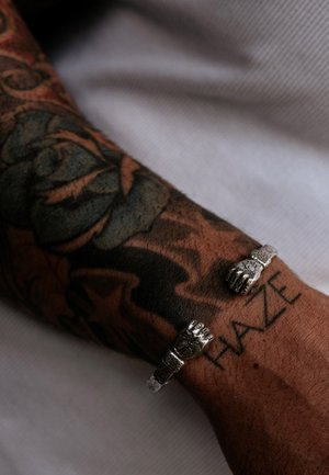 Sølv cuff-armbåndet har to dekorative hænder og intrikate detaljer, båret på en tatoveret underarm med en synlig "HAZE" tatovering.