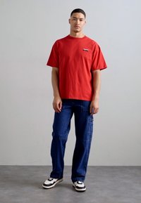 Rød kortærmet bomulds T-shirt med lille logo på brystet, parret med løse blå denim cargo bukser og sort-hvide sneakers.