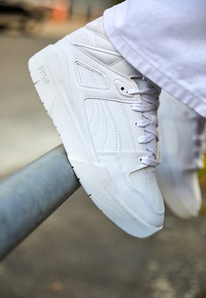 Biały sportowy trzewik na sznurowadła spoczywający na metalowej poręczy, noszony z białymi spodniami, rozmyte tło na zewnątrz.