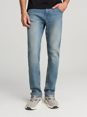 Jeans di denim azzurro chiaro con vestibilità slim, caratterizzati da cinque tasche e una sottile texture sbiadita. Indossati con scarpe da ginnastica grigie.