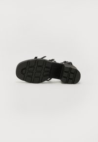 Black sandal with a textured rubber sole, featuring a flat heel, multiple straps, and an adjustable buckle for closure.