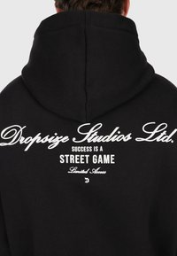 Personne portant un sweat à capuche noir avec un texte blanc au dos qui dit "Dropsize Studios Ltd. Le succès est un jeu de rue accès limité."