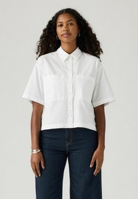 White short-sleeve cropped shirt with a button-down front and two chest pockets. Smooth cotton fabric, relaxed fit, minimal design accents.