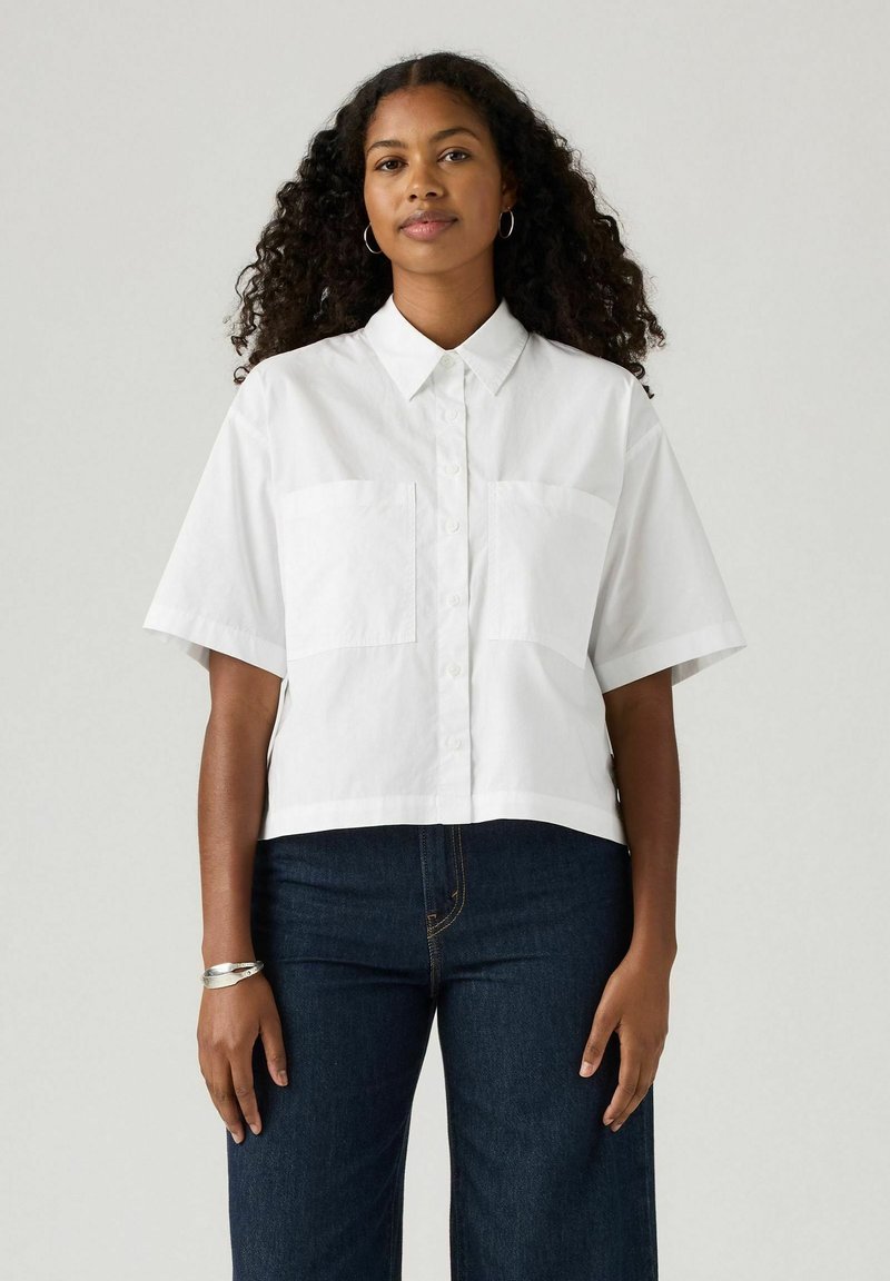 White short-sleeve cropped shirt with a button-down front and two chest pockets. Smooth cotton fabric, relaxed fit, minimal design accents.
