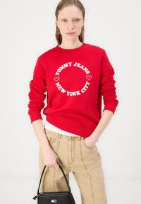 Rød hettegenser med hvit sirkulær logo "TOMMY JEANS NEW YORK CITY". Bomullsblanding, ribbestrikkede mansjetter og hem. Kombinert med beige bukser og en svart håndveske.