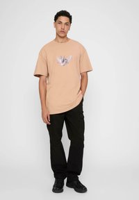 Lys beige oversized t-skjorte med grafikk av en fugl på fronten; sammen med svarte cargobukser og svarte joggesko. Enkelt bakgrunn.