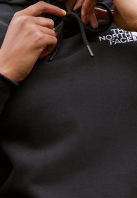 Svart huvtröja med dragsnören, med en vit broderad logotyp på bröstet. Mjuk tygkänsla, avslappnad passform.