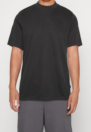 Osoba ubrana w jednokolorowy czarny krótki rękaw i szare szorty, stojąca na tle jednolitego białego tła.