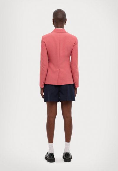 Blazer in light pink fabric with structured design, single vent at the back, paired with navy shorts and black shoes.