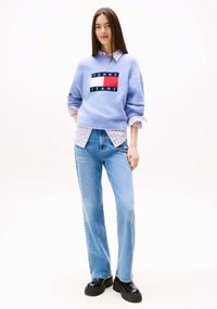 Maglione blu lavorato a maglia con logo "TOMMY JEANS", indossato sopra una camicia a quadri chiara. Un paio di jeans dritti azzurri e scarpe nere chunky.