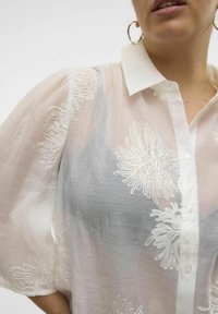 Weißes, halbtransparentes Hemd mit Puffärmeln, das zarte weiße Blumenstickereien und Knöpfe auf der Vorderseite zeigt. Strukturierter Stoff, weicher Fall.