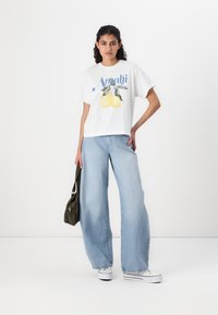 Hvid cropped t-shirt med citron grafik og blå tekst, parret med lyseblå brede jeans og hvide sneakers. Olivengrøn taske holdt.