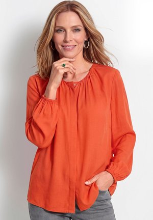 Femme aux cheveux blonds mi-longs porte des boucles d’oreilles anneaux et un chemisier orange à manches longues, souriante, une main près du visage et l’autre dans la poche.