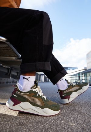 Persoon draagt olijfgroene, witte en bordeauxrode sneakers en witte Puma-sokken, zittend op een metalen bankje buitenshuis op een zonnige dag.