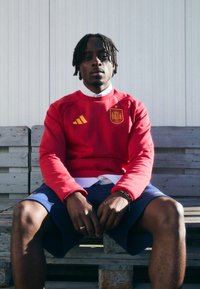 Sarkans džemperis ar dzelteno Adidas logo un Spānijas �ģerboni, kombinēts ar tumšām biksēm. Audumam izskatās gluda tekstūra, un piegriezums ir brīvs.