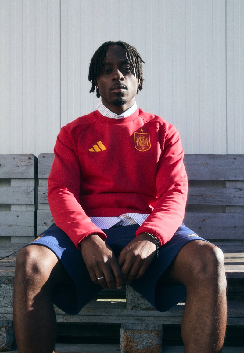 Červená mikina s kapucí a žlutým logem Adidas a španělským erbem, doplněná tmavými šortkami. Textura vypadá hladce a střih je volný.