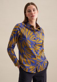 Camicetta blu e gialla con motivo a fantasia, caratterizzata da un design con bottoni, maniche lunghe e una texture liscia con motivi di foglie ondulate.